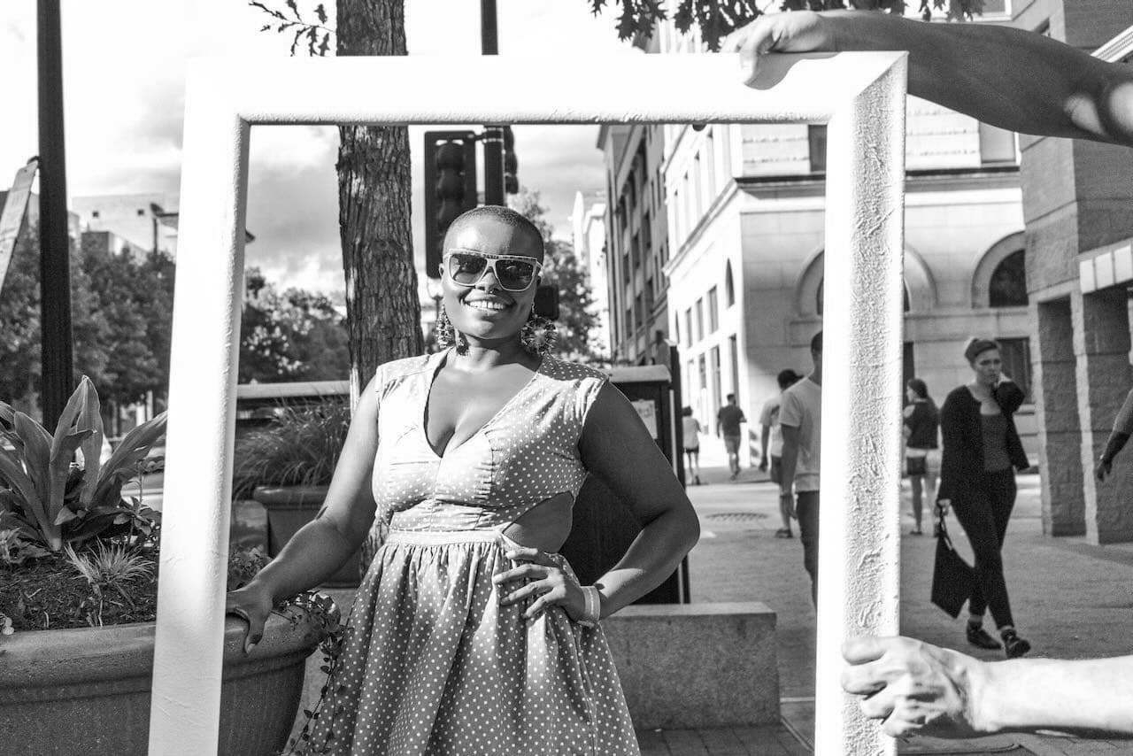 A smiling woman wearing sunglasses and a polka-dot dress poses behind a picture frame held by others on a busy city sidewalk with buildings and pedestrians in the background.