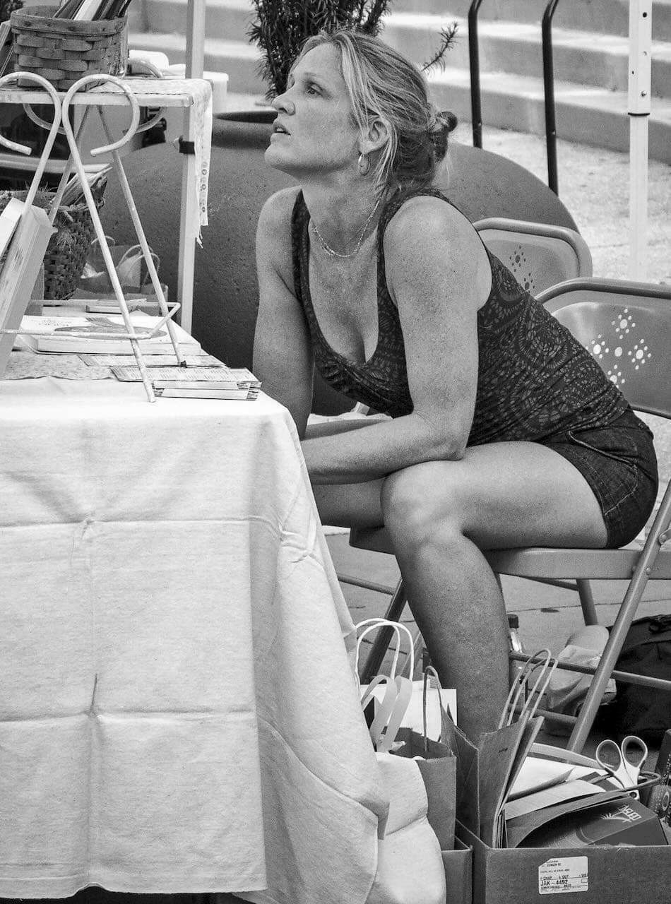 A woman sits on a foldable chair, resting her arms on her knees and looking intently forward; she is among tables covered with books, boxes, and bags in an outdoor market.