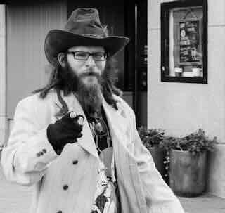 Bearded man wearing a hat and glasses gesturing with a finger while walking on a sidewalk in an urban area with a building and potted plants in the background.