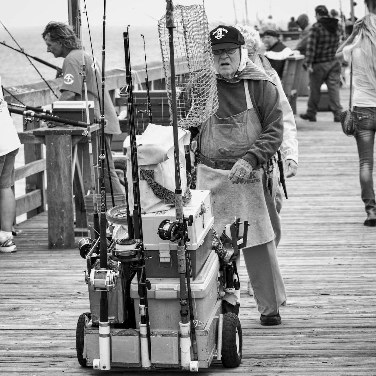 An elderly man pushes a cart full of fishing rods and equipment on a busy wooden pier crowded with people engaged in various activities.