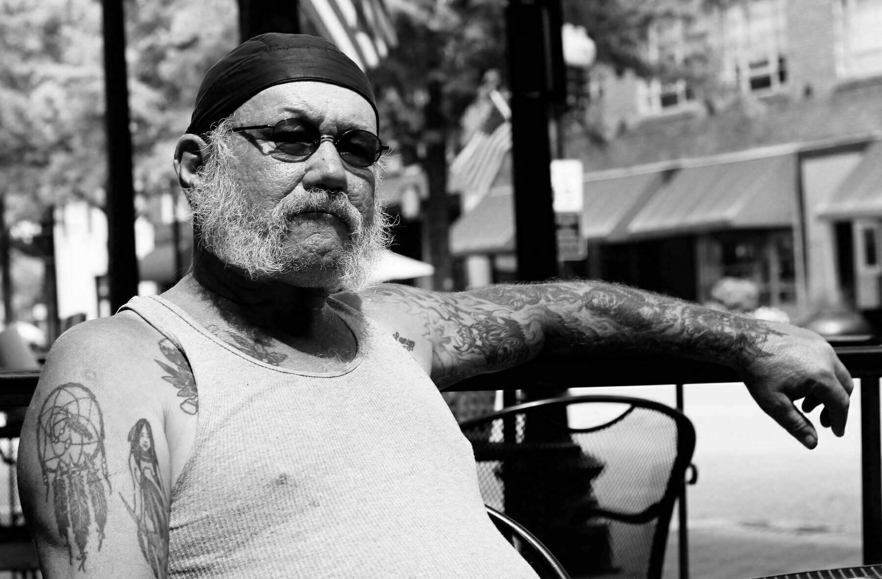 Man with tattoos sits outdoors, wearing a tank top and bandana, arm resting on chair, in a shaded area of a street with storefronts and trees.