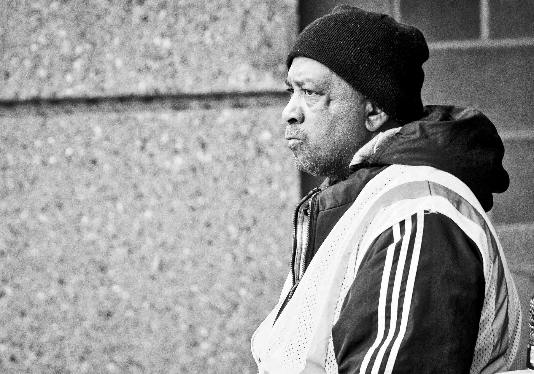 A man wearing a knit hat and striped jacket stands in profile, looking serious, against a textured concrete wall.