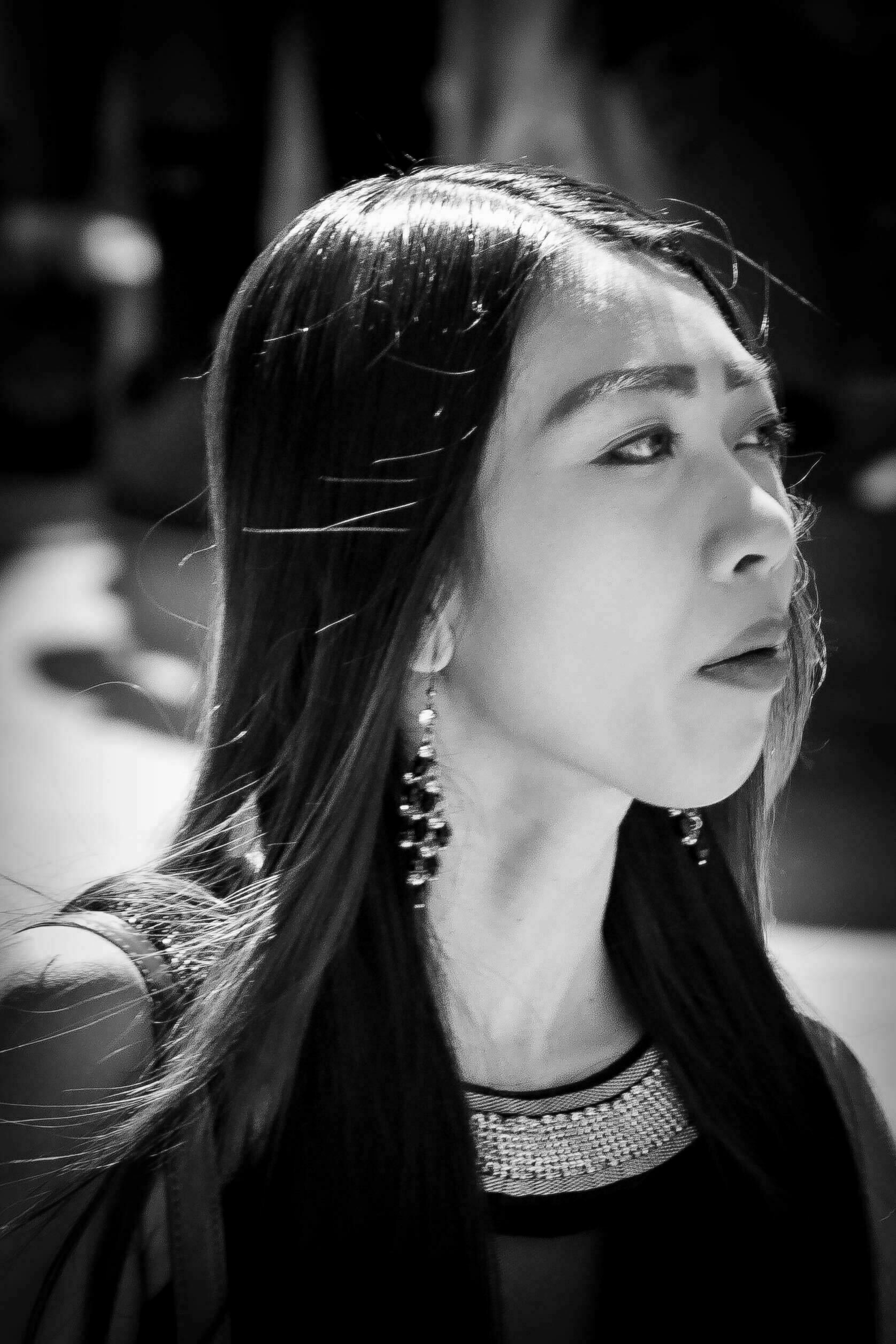 Close-up of a woman with long, straight hair, wearing earrings, looking slightly upwards, in a black and white setting with other indistinct figures in the blurred background.