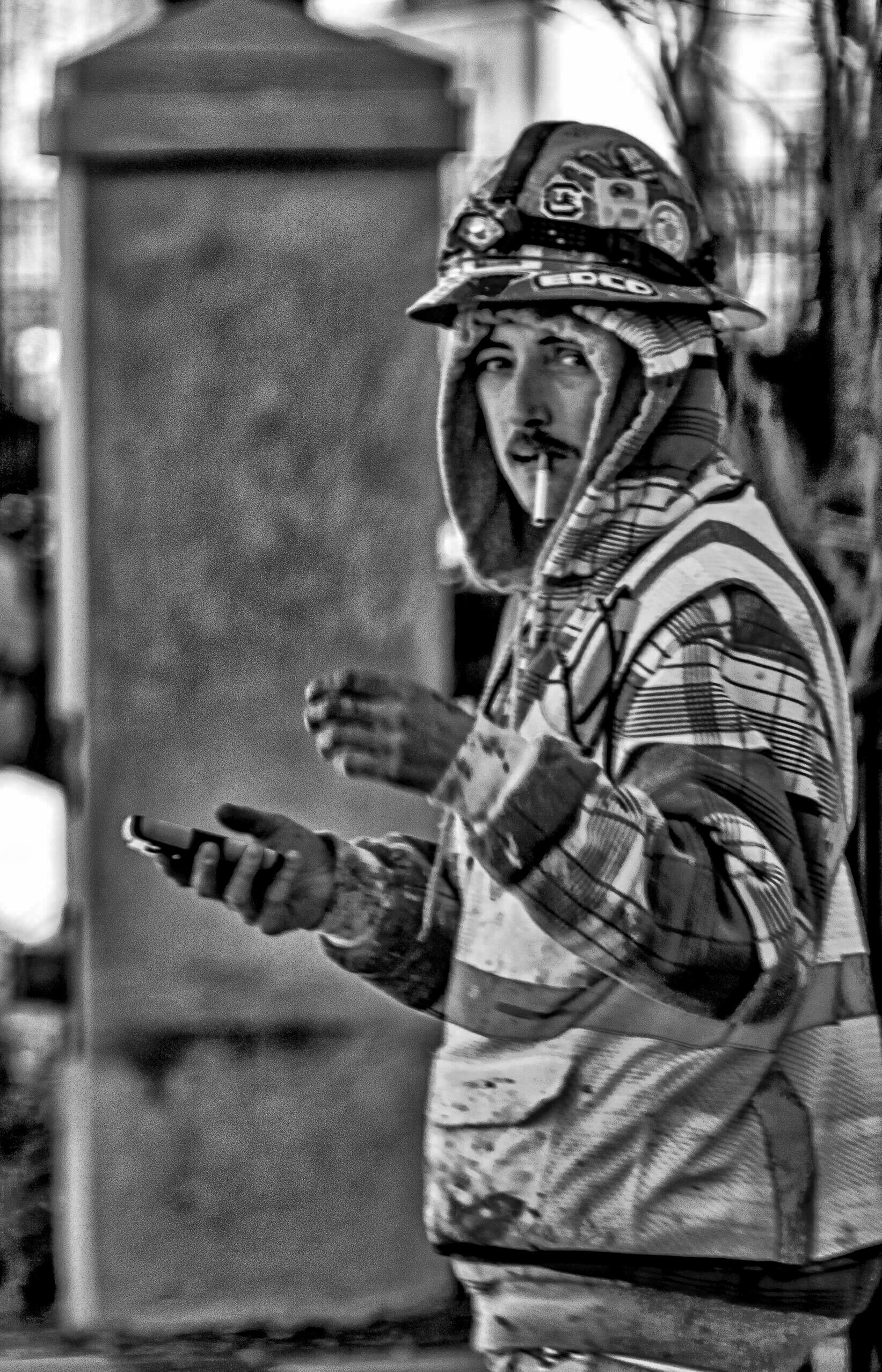 A construction worker wearing a hard hat and a hooded jacket holds a smartphone in one hand and a cigarette in their mouth, standing outdoors near a concrete pillar.