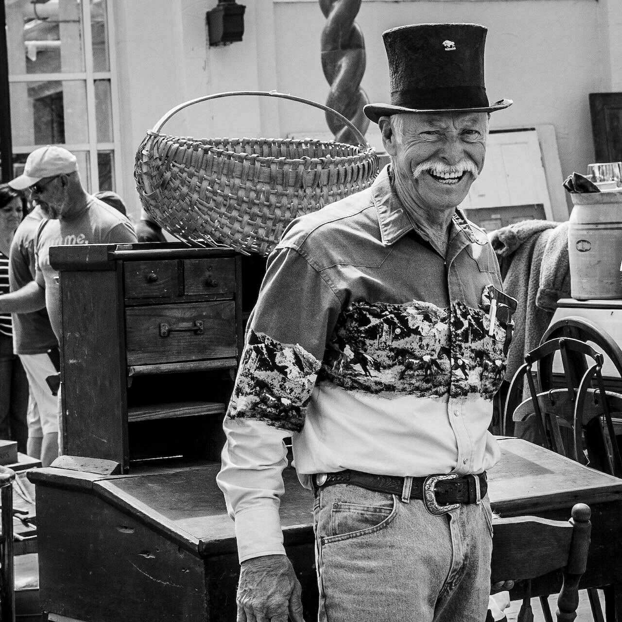 Man smiling at an outdoor antique market against a background of old furniture and people browsing.