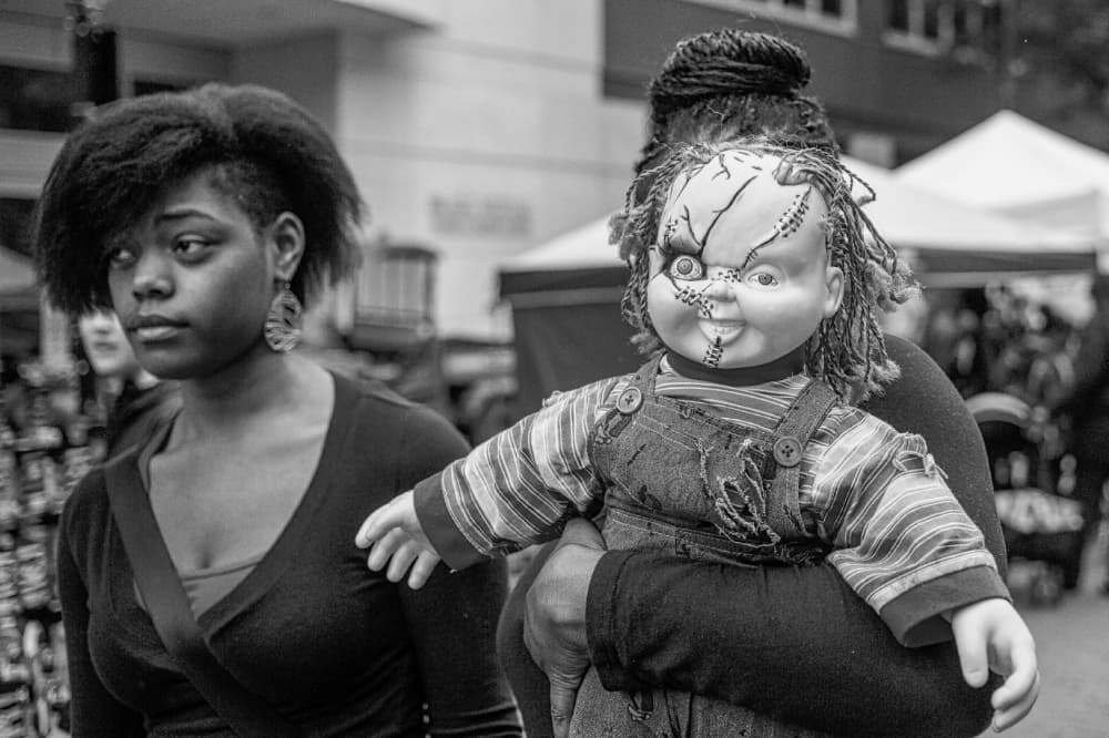 A person carries a large stitched-up, dreadlocked doll with a creepy smile while another person looks ahead in an outdoor market setting.