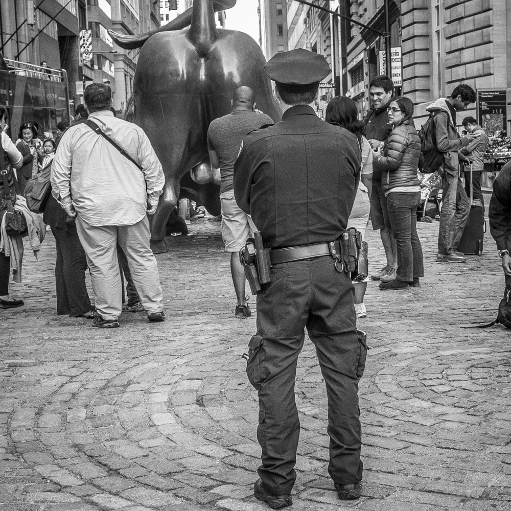 Rear view of the famous bull statue on Wall Street in NYC surrounded by tourists supervised by a police officer