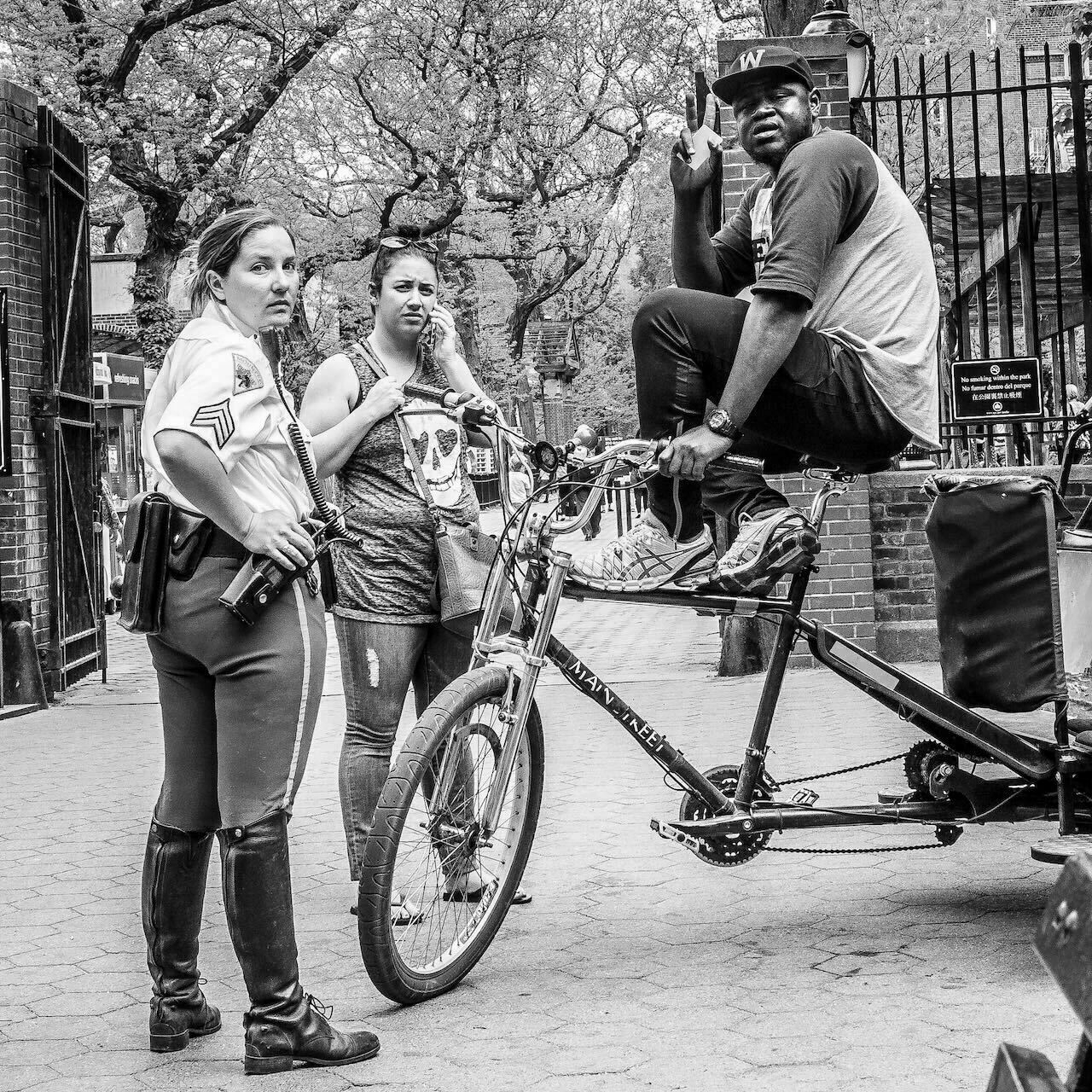 A police officer and a woman observe a man giving a peace sign while sitting on a rickshaw in a tree-lined park area.