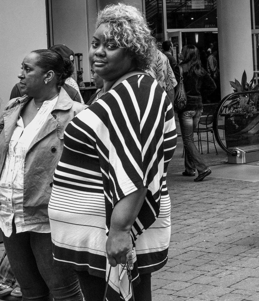 Woman with curly hair wearing a striped top looking at the camera, standing with another woman in an urban area with people and an escalator in the background.