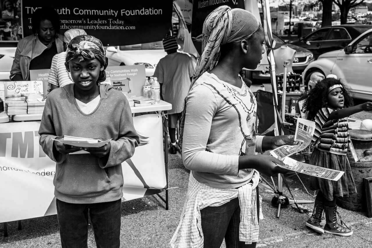 Three children hold flyers, standing in front of a booth with the “TMF Youth Community Foundation” banner, with books and informational materials on display. Cars and people fill the background.eg