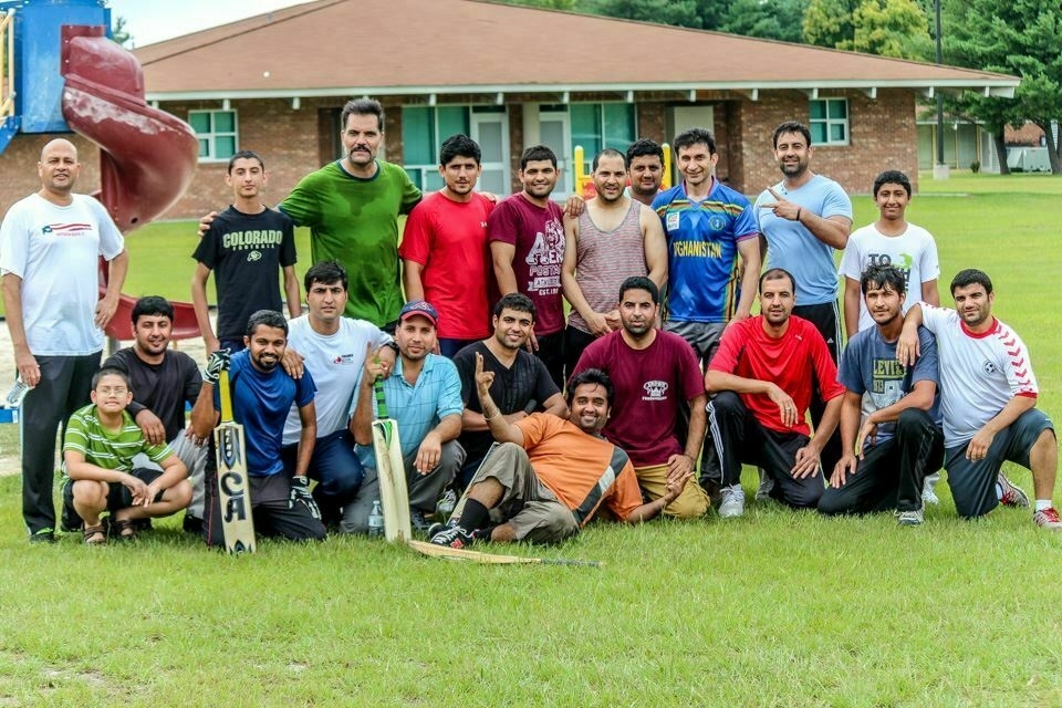 A group of 22 men and boys are posing together outdoors, some holding cricket equipment, in front of a brick building with a playground slide visible in the background.