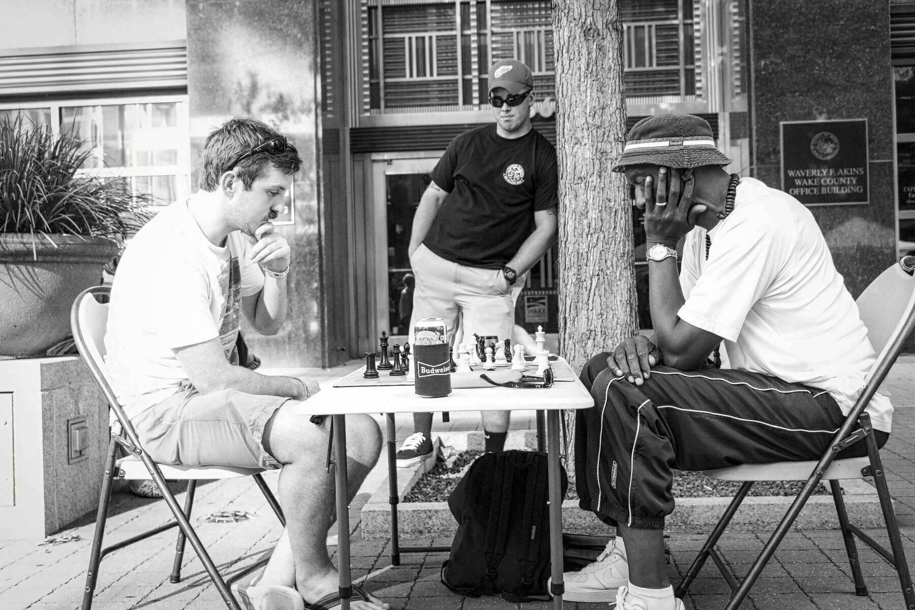 Two men, sitting across from each other, are intensely playing chess outdoors. A third man is standing and watching them. The backdrop includes urban street elements and the Waverly F. Akins Wake County Office Building sign.