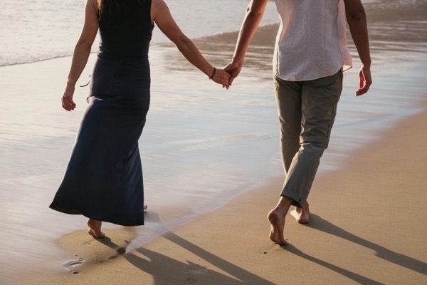 A couple walking down the beach holding hands shown from the shoulders down in the back
