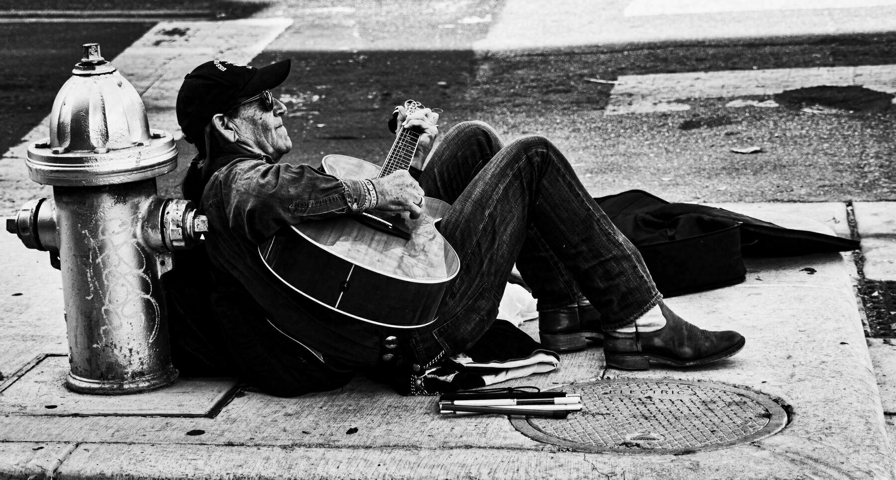 A person rests against a fire hydrant, playing a guitar, seated on a sidewalk, with a guitar case and walking cane for the blind  nearby. Urban street setting.