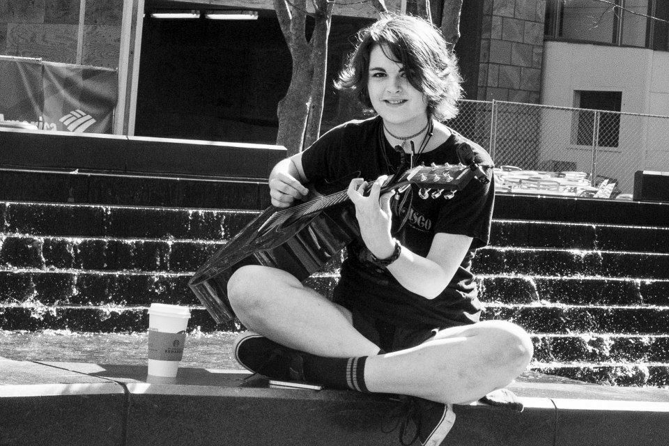 A young female busker playing the guitar while sitting cross-legged beside a coffee cup