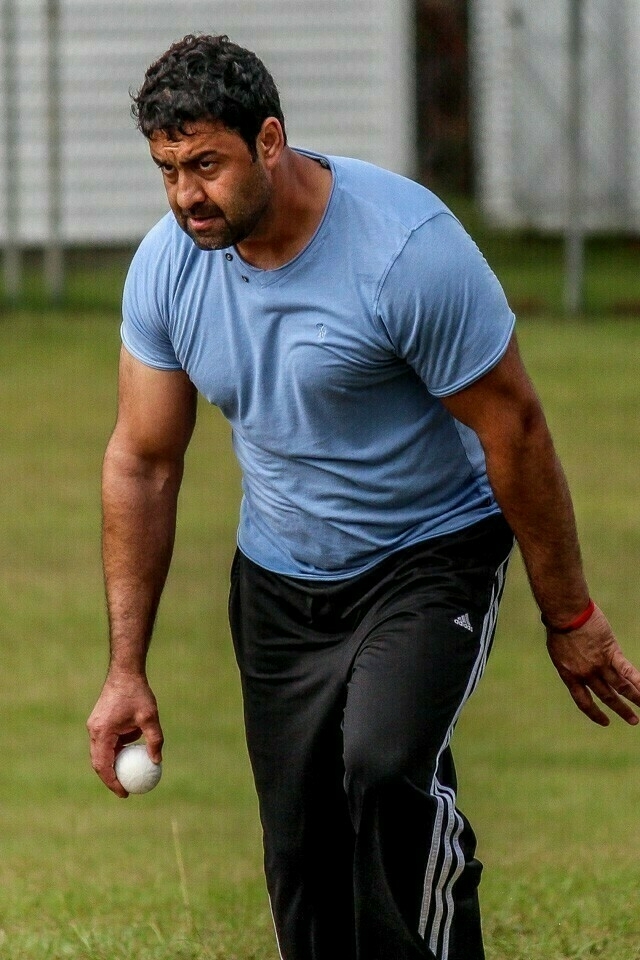 A man in a light blue shirt and black track pants is holding a white ball and appears to be in mid-action on a grassy field.