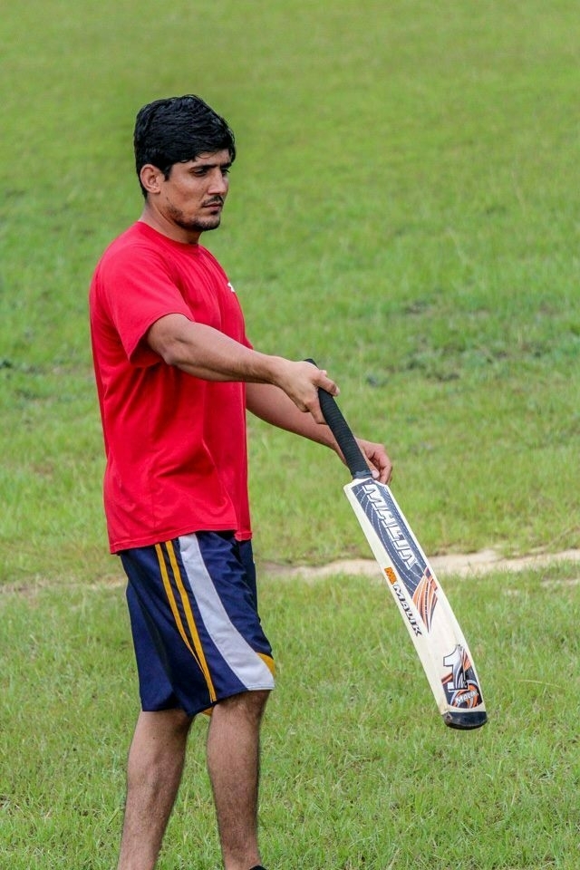 A young man in a red shirt and navy shorts is holding a cricket bat on a grassy field.