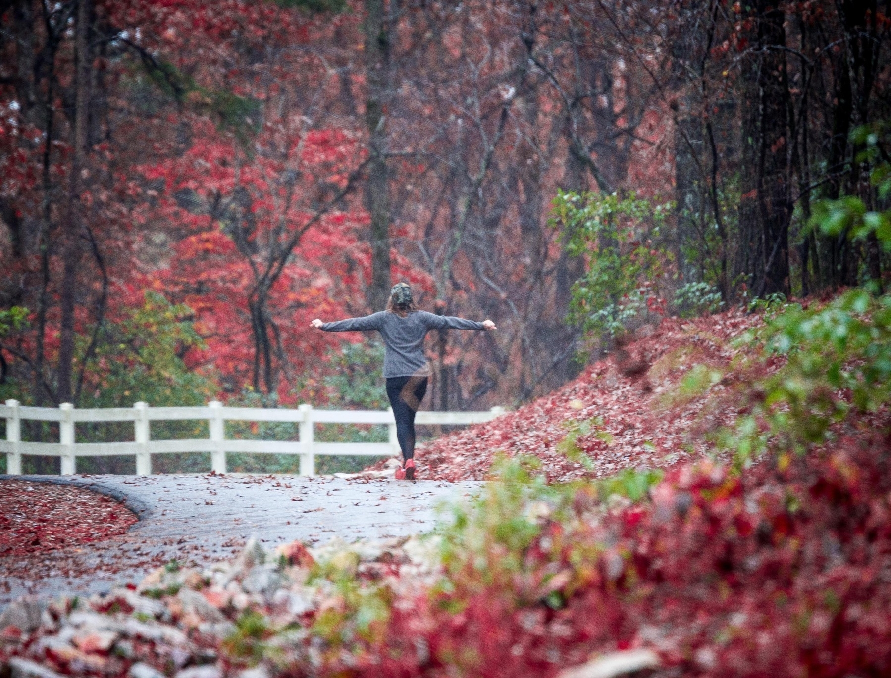 A woman with arms extended walks along an asphalt path in woods with autumnal colors.