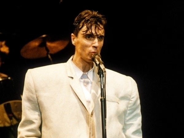 [David Byrnes in the white suit he wore for Stop Making Sense singing into a microphone]