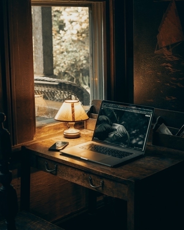 A Macbook and an iPhone sitting on a wooden desk lit by a table lamp