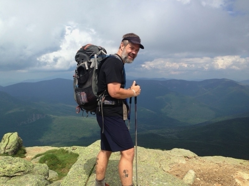 Auto-generated description: A man with a backpack stands on a rocky mountain peak, holding a hiking pole and smiling with a panoramic view of distant mountains and cloudy sky behind him.