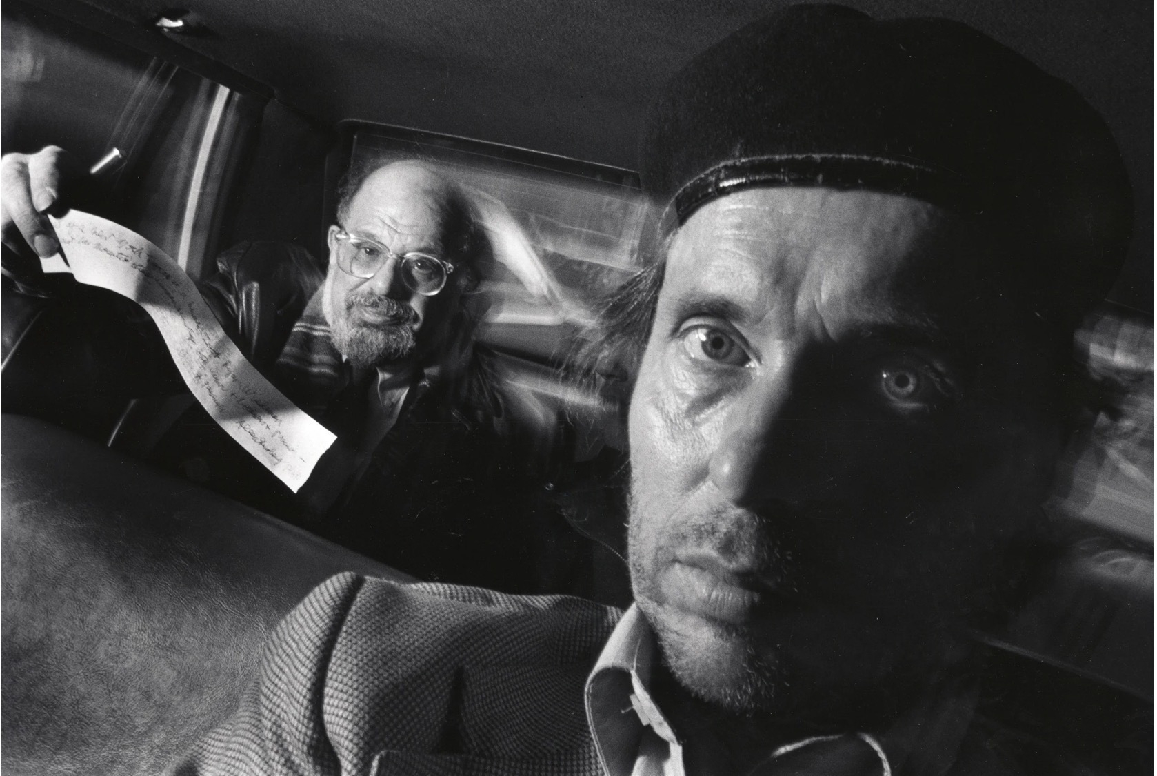 Two men are inside a car, Alan Ginsberg holding a piece of paper while the other looks directly into the camera.