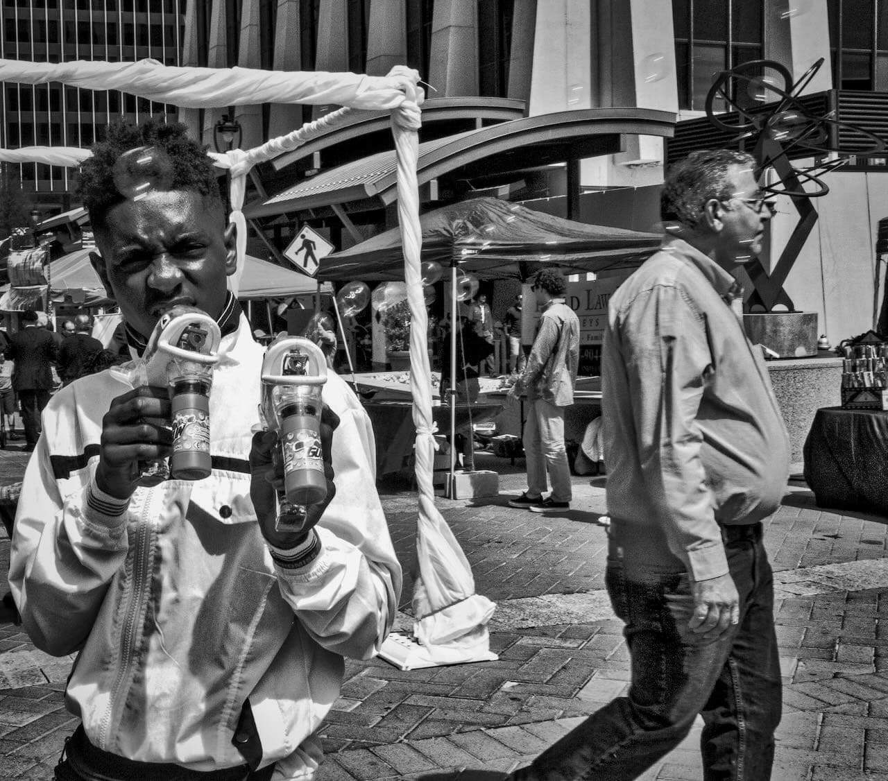 Person holding bubble dispensers, standing in an outdoor market; people walk and browse vendor stalls under tents amidst urban architecture.