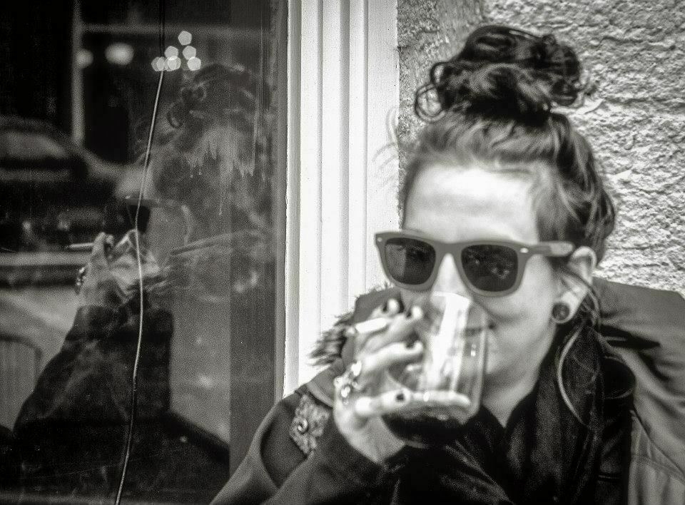 A woman outside taking a drink from a glass while holding a cigarette with the same hand