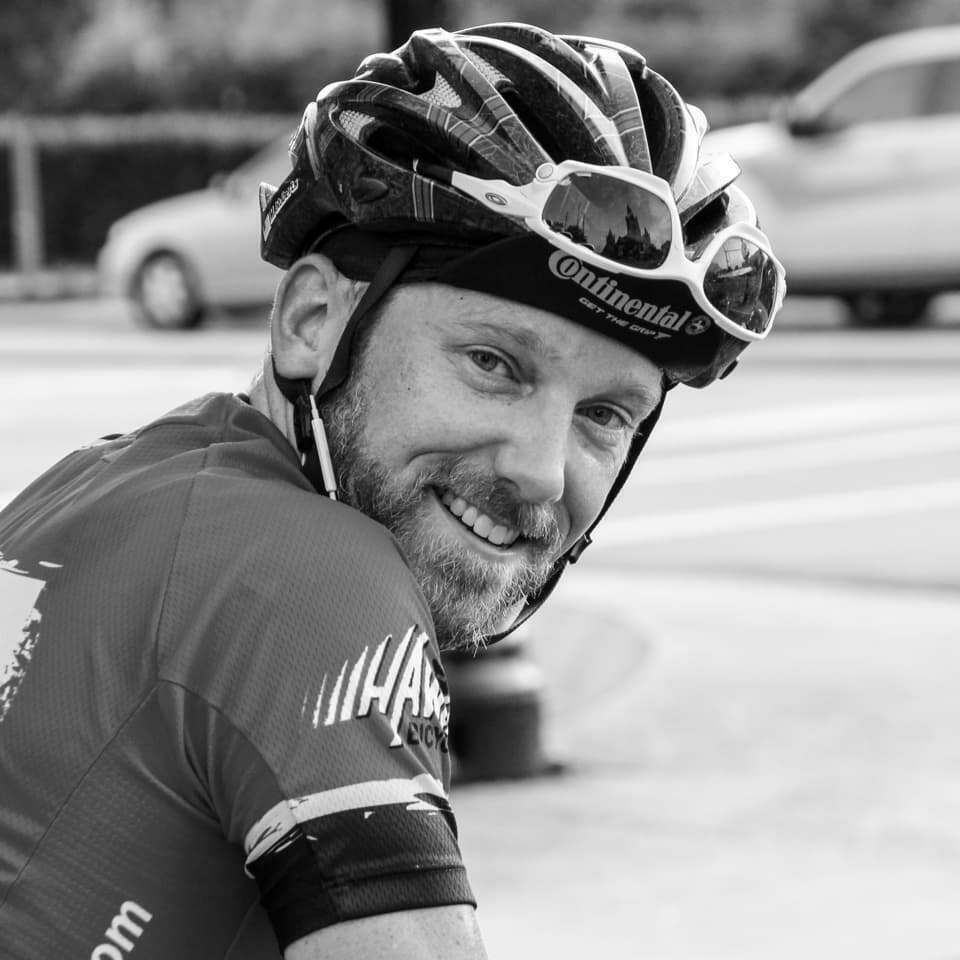 A cyclist with a beard smiles while wearing a helmet and sunglasses, situated outdoors near a street with cars in the background.