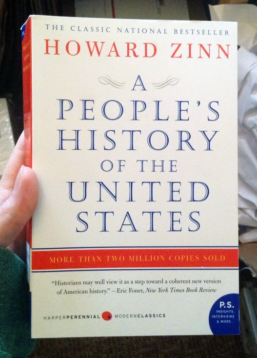 The red, white and blue book cover of People's History of the United States by Howard Zinn, held in a pair of hands