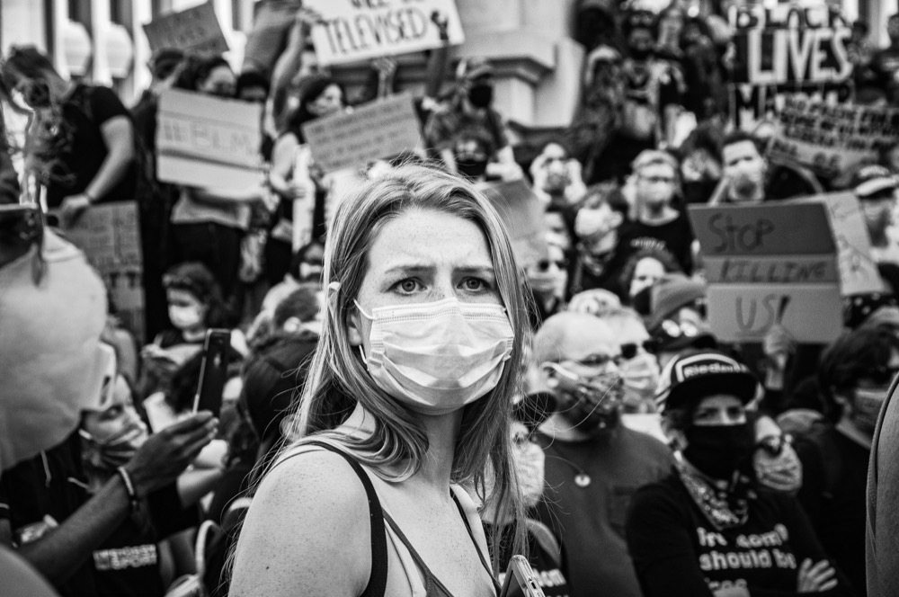 A black and white image of a woman wearing a breathing mask and a tank top surrounded by sign carrying protesters