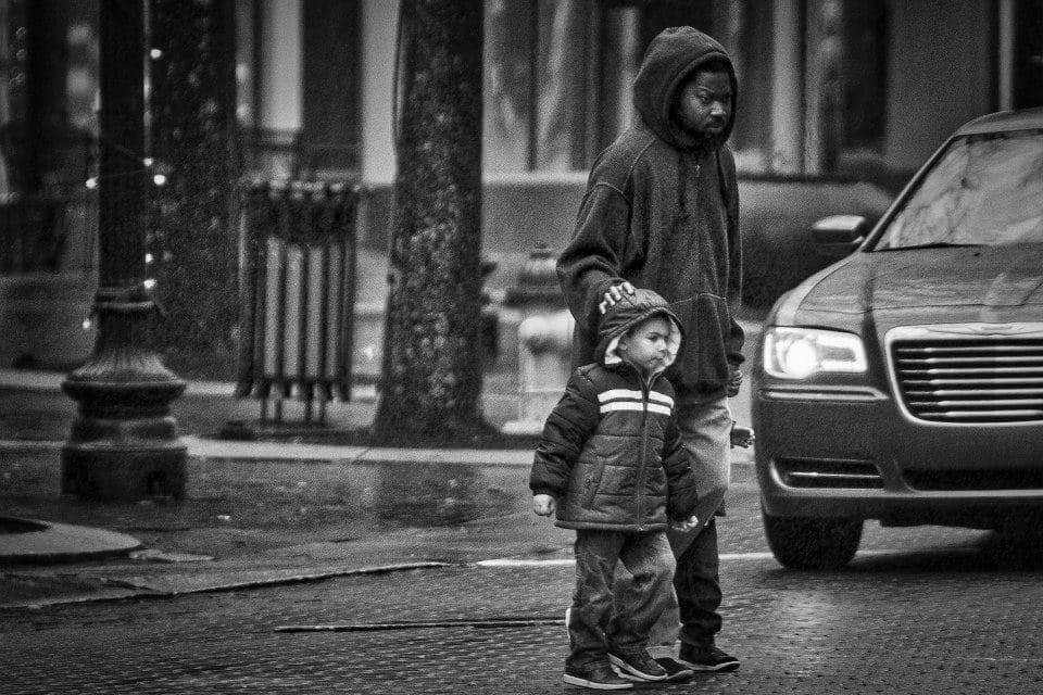 An adult in a hoodie guides two young children in coats, walking across a wet street with a parked car and city buildings in the background.