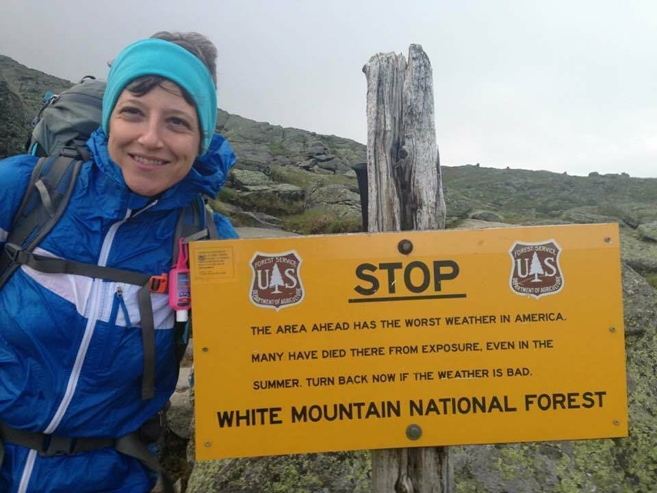 A smiling person dressed in a blue jacket and wearing a headband stands next to a warning sign from the White Mountain National Forest that cautions about the area's bad weather and the risks of exposure.