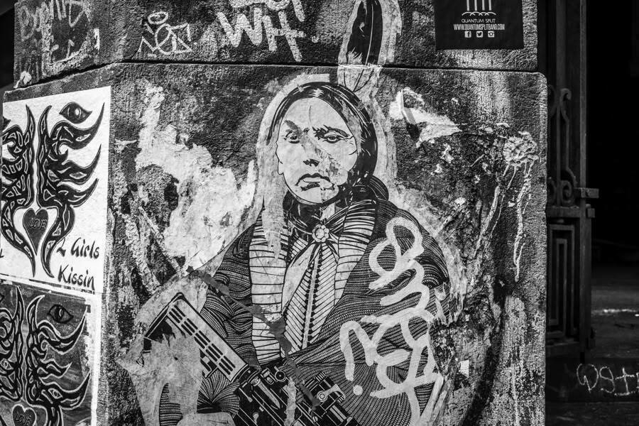 A black and white mural on a textured wall depicts a person in traditional attire holding a rifle. The image is surrounded by graffiti and street art, including stylized eyes and a heart with the word "LOVE" written on it. The wall has various layers of textures, paint, and other artworks, contributing to a gritty urban atmosphere.