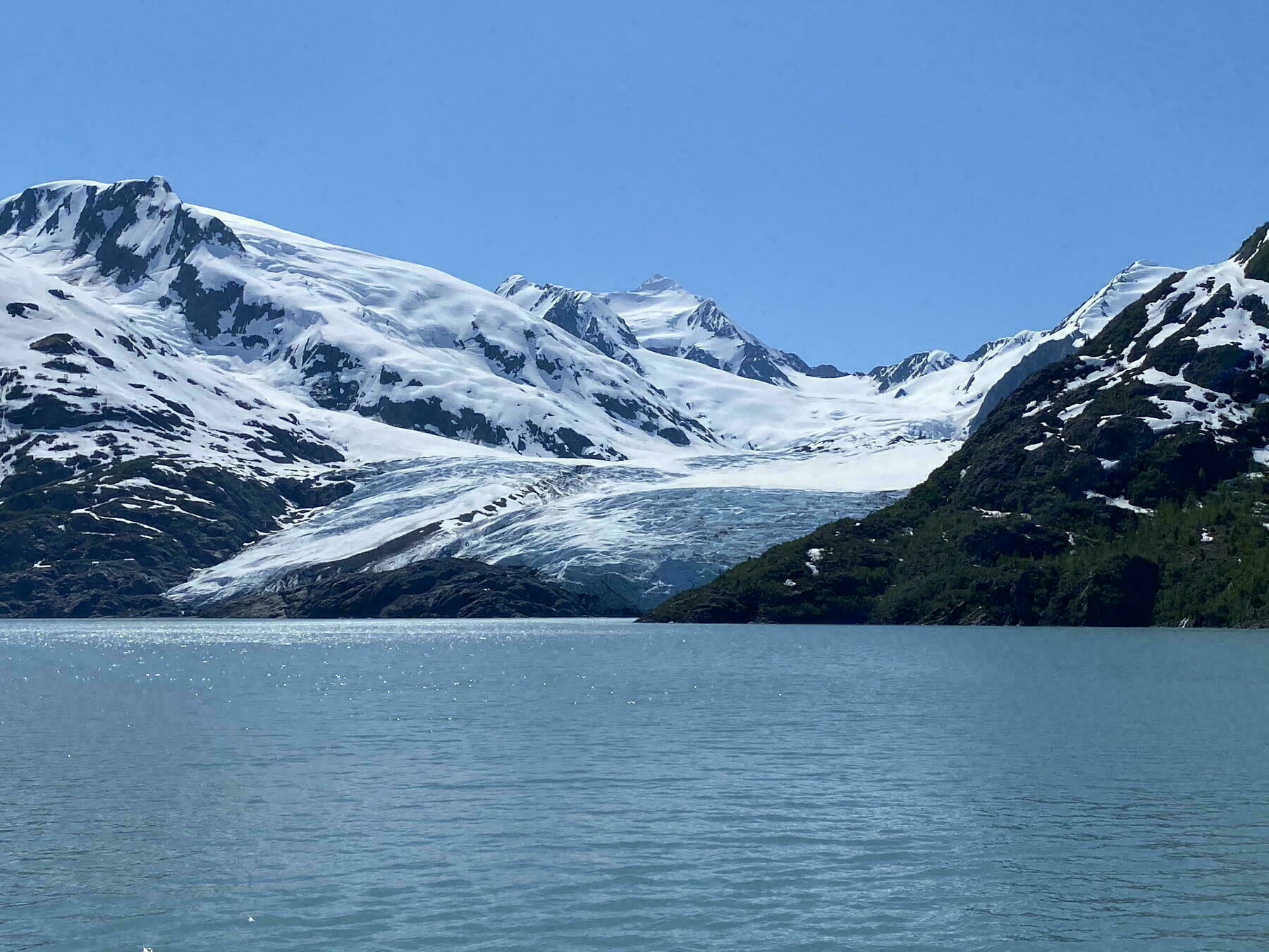Snow-covered mountains surround a glacier flowing between rocky slopes, meeting a calm, turquoise lake under a clear blue sky.