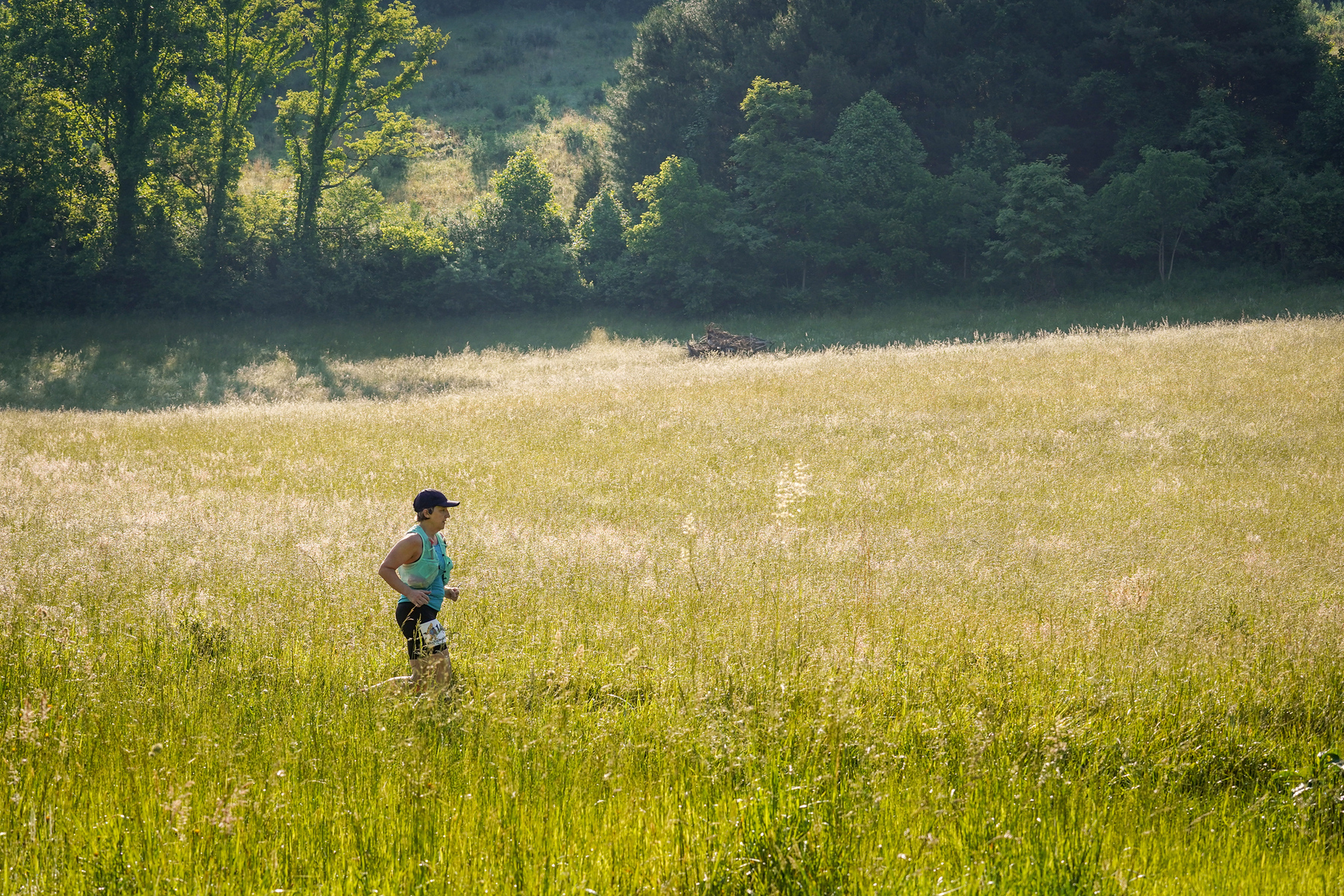 A person is running through a sunlit grassy field surrounded by trees.