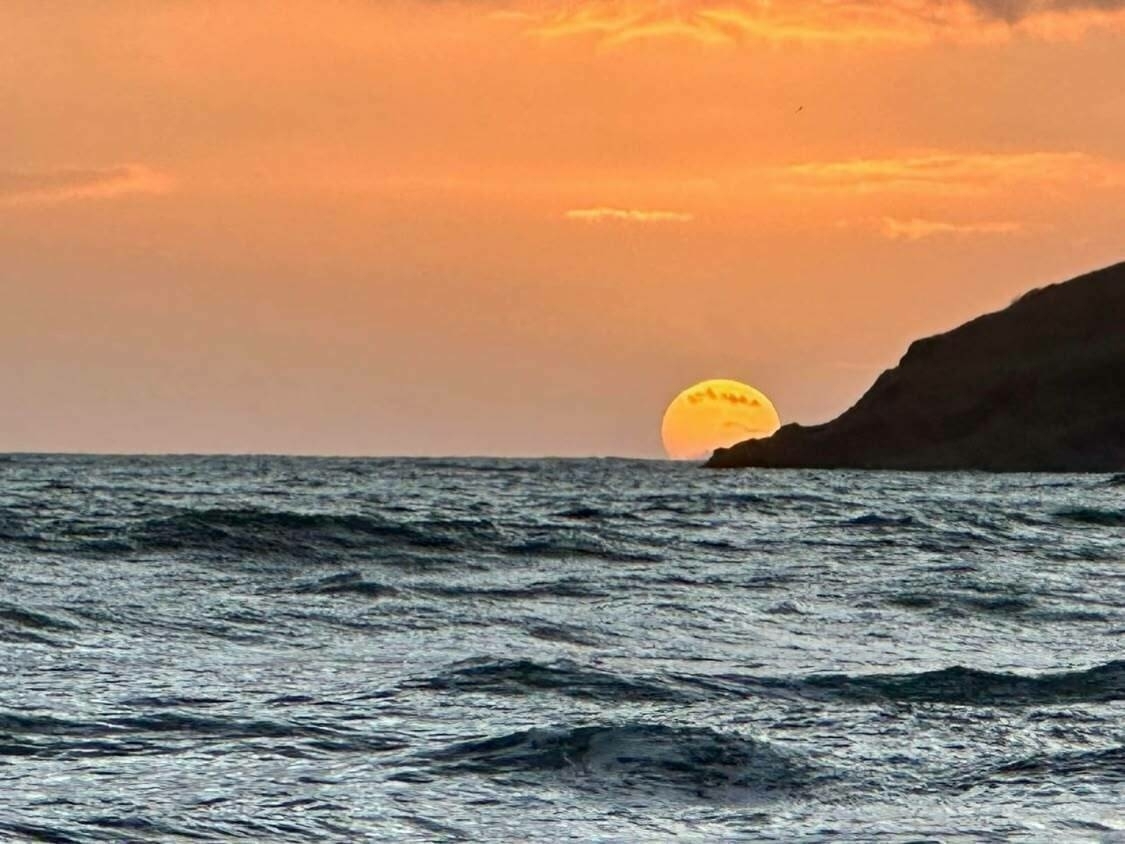 Sun setting partially beyond a rocky shoreline, casting a warm orange glow over the ocean waves in a tranquil seaside scene.