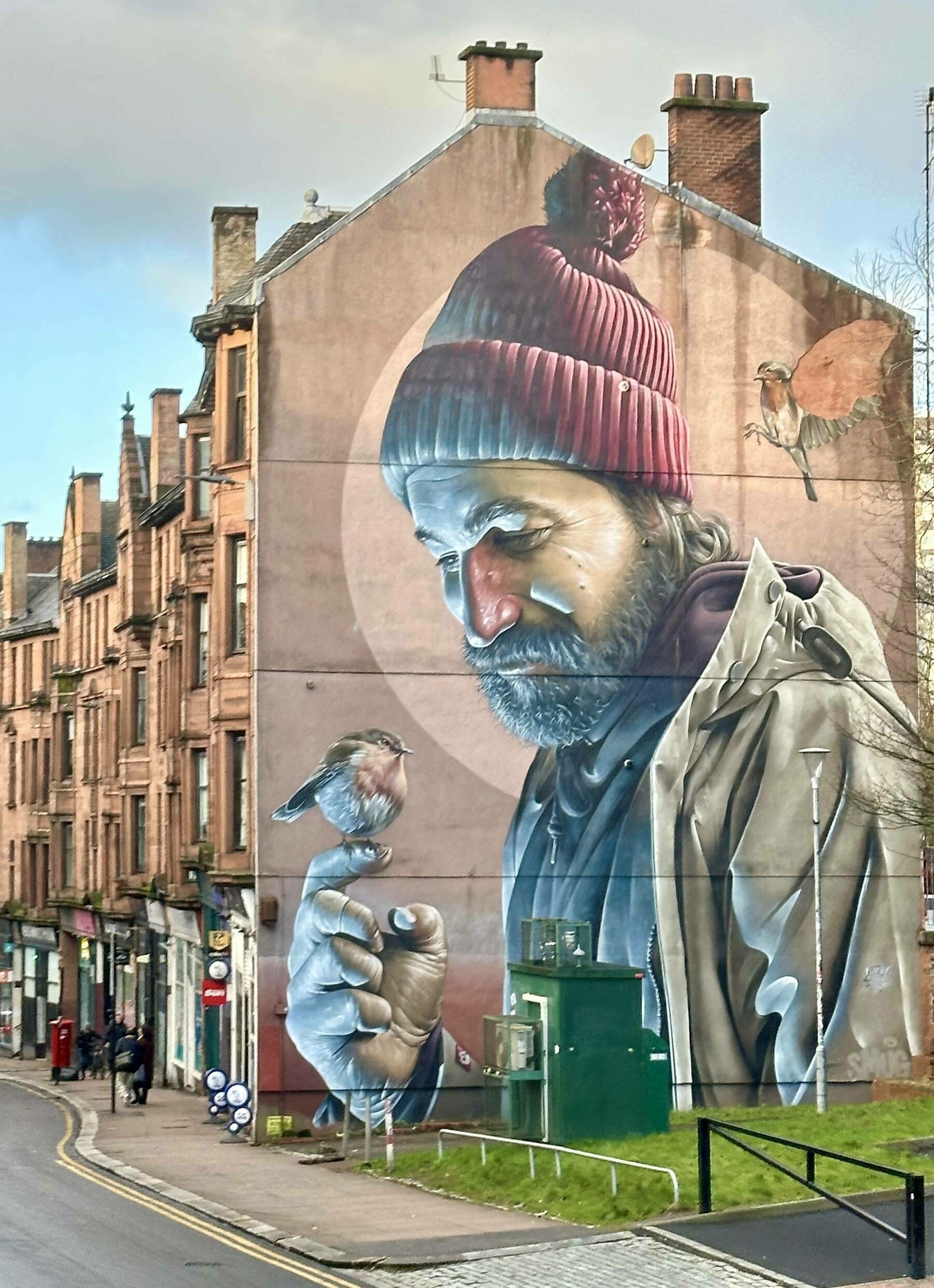 Mural depicts a bearded man with a red beanie, gently holding a small bird on his finger, set against a large city building. Nearby, old brick buildings line the street.