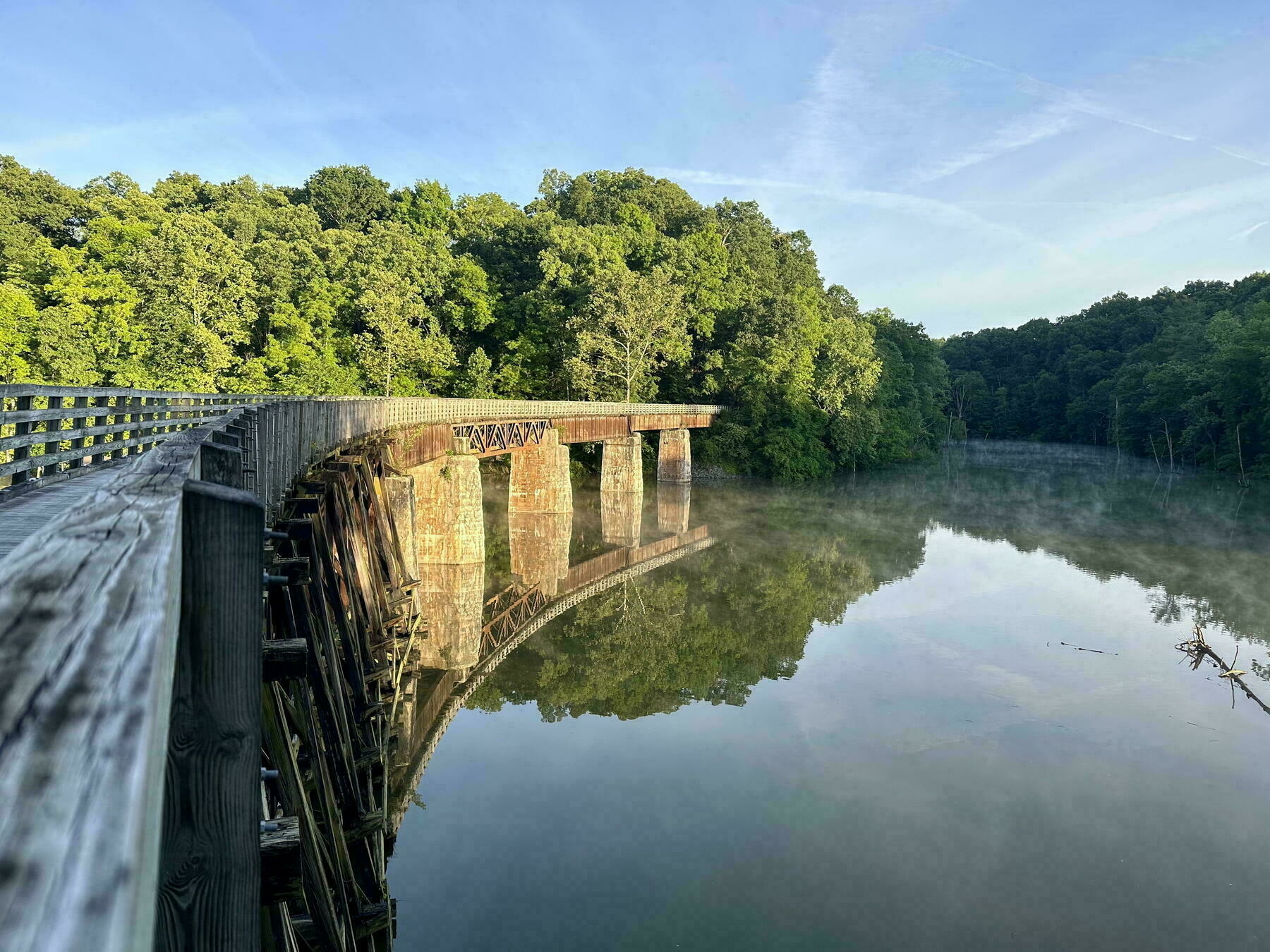 A wooden bridge curves over a serene, mist-covered river surrounded by lush green trees.