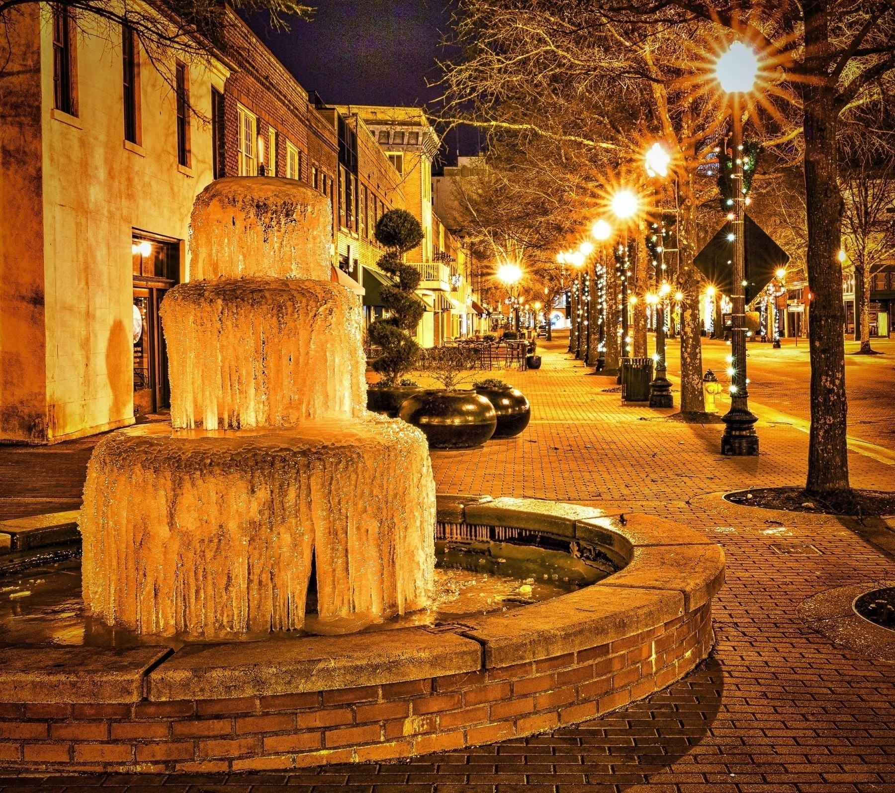 A frozen fountain is illuminated by streetlights along a brick-paved pedestrian walkway lined with trees at night.