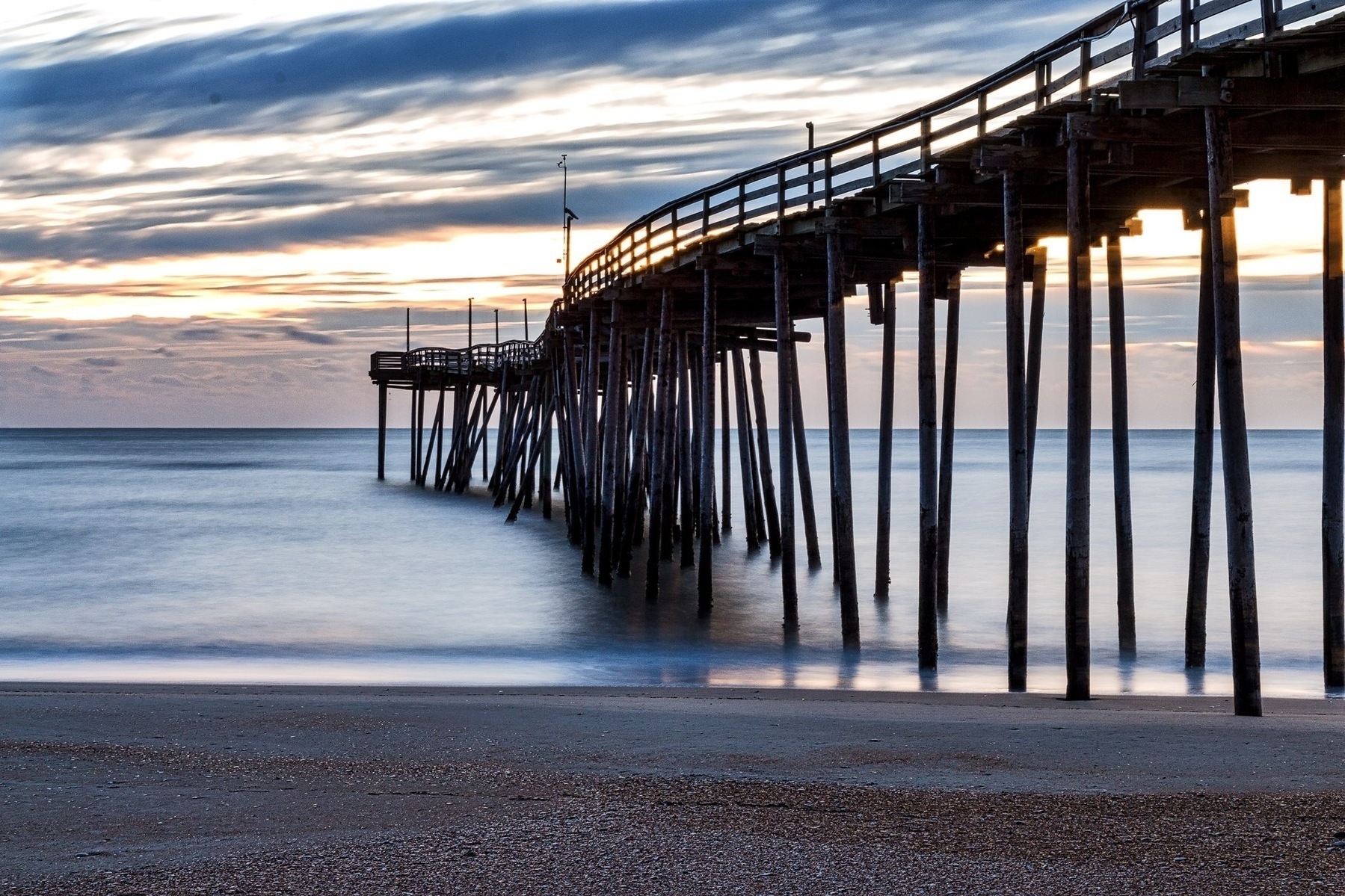 A wooden pier extends over calm ocean waves; the sunset sky glows with orange and blue hues, creating a serene coastal atmosphere.