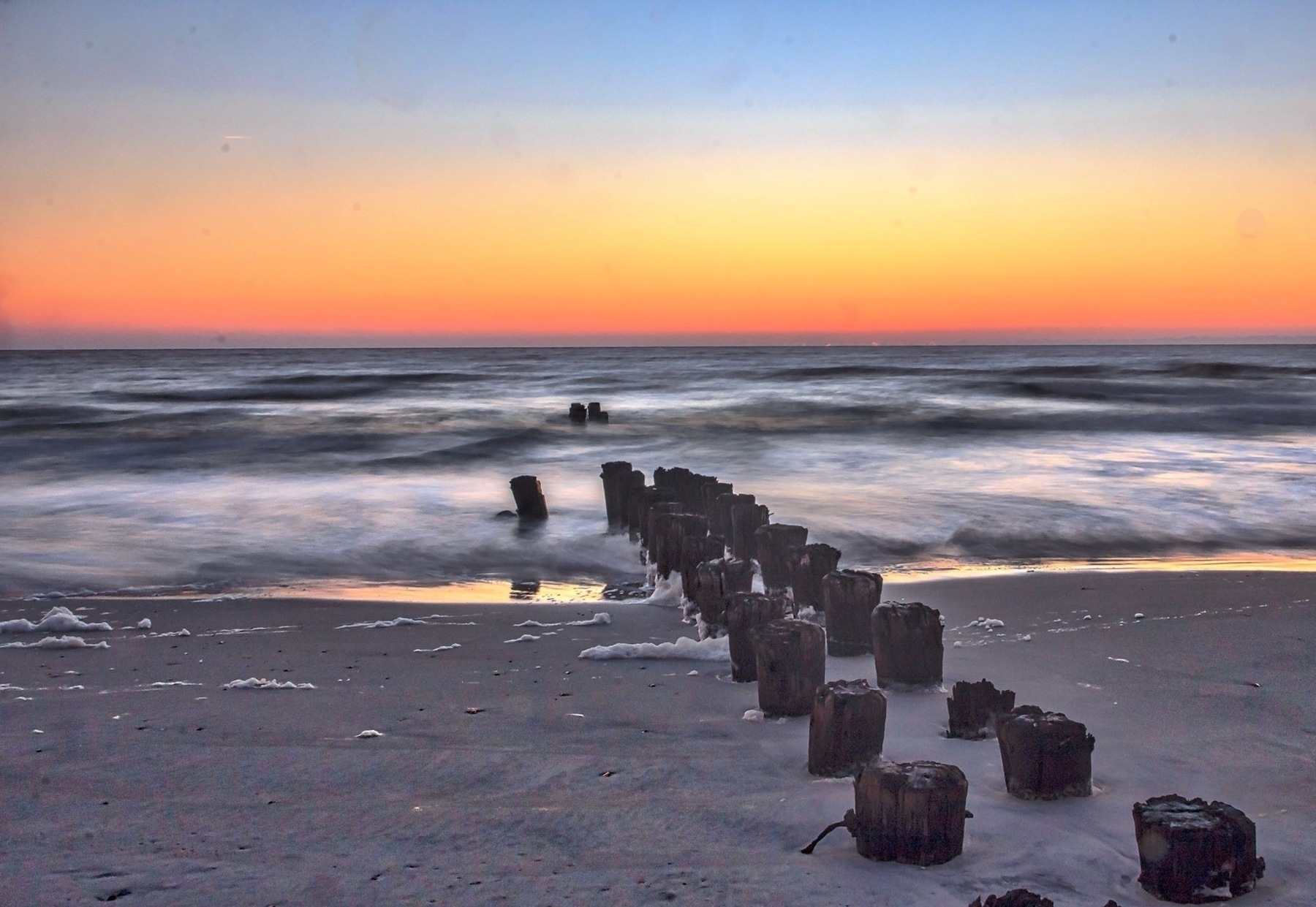 A serene sunrise over a beach features wooden groynes extending into the calm ocean waves.