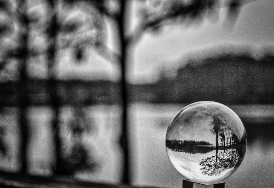 A crystal ball in focus shows winter trees reflected in a lake  - in black and white.