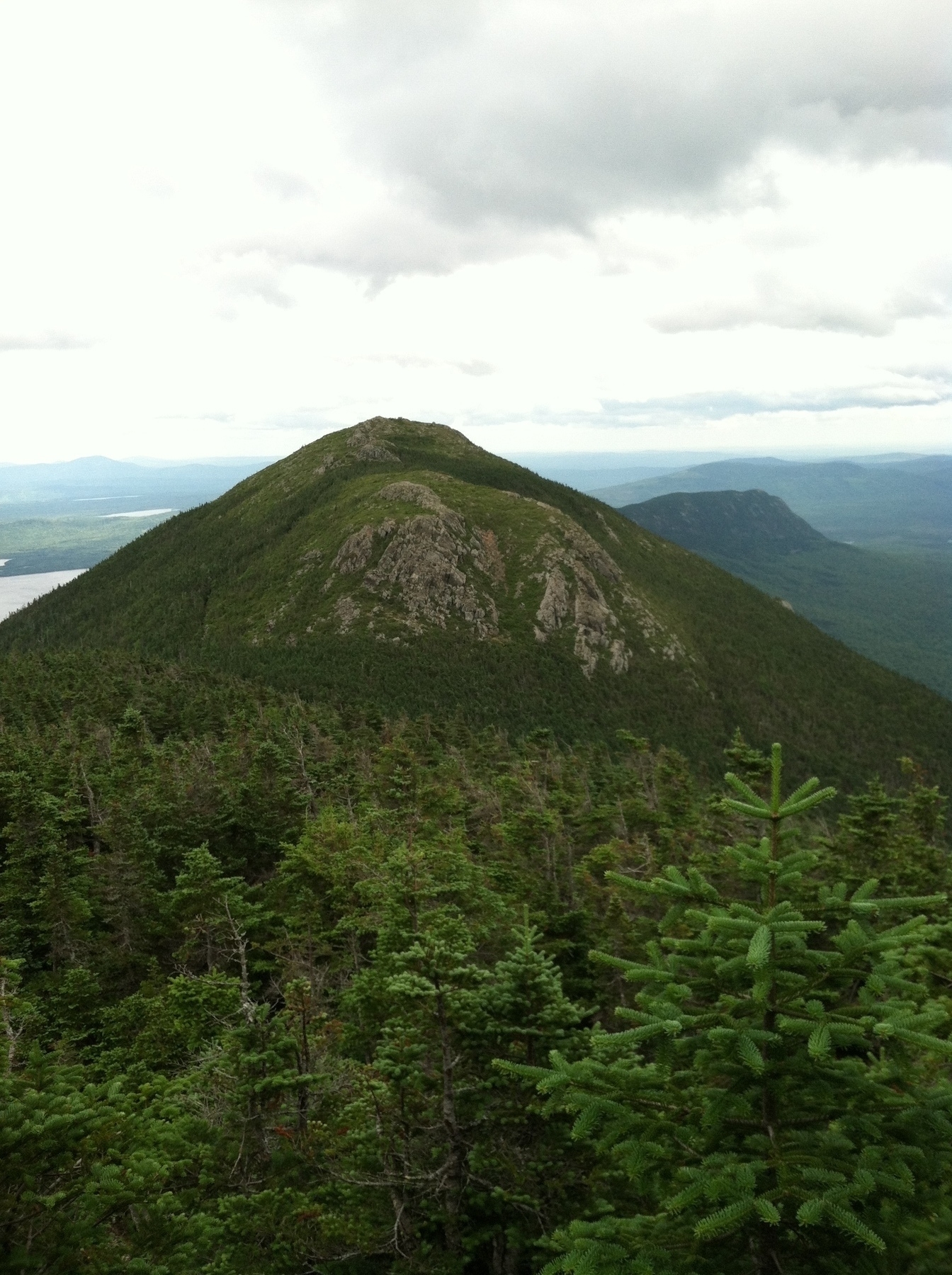 A vast landscape features a lush green forest foreground, scattered lakes, and a dramatic cloudy sky above distant mountains.