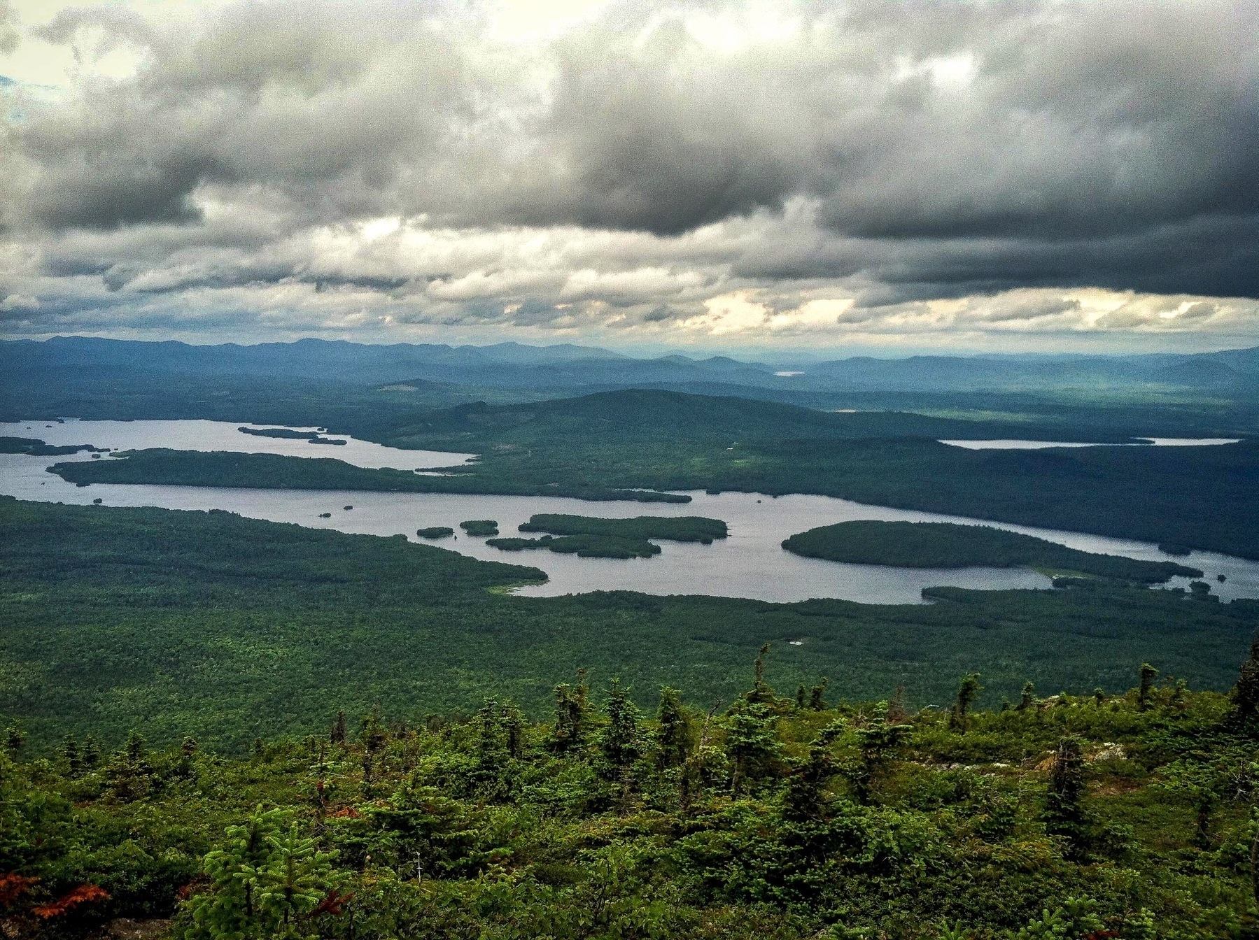 A vast landscape features a lush green forest foreground, scattered lakes, and a dramatic cloudy sky above distant mountains.