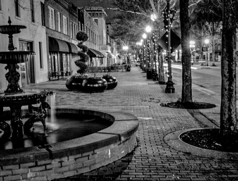 Fountain with water cascading sits in foreground, while trees and streetlights line a deserted sidewalk beside buildings in a nighttime urban setting.