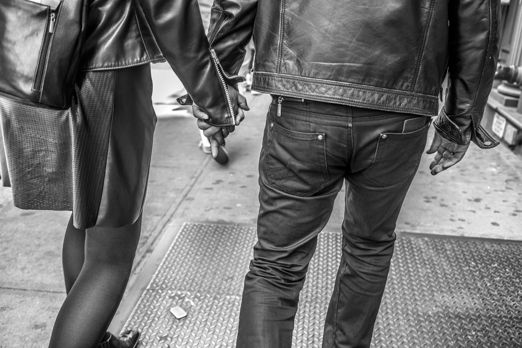 A black and white photograph showing two people walking hand in hand on a city sidewalk. Both are wearing leather jackets and dark pants. The person on the left is carrying a backpack and wearing a skirt with opaque tights, while the person on the right is in jeans. Their faces are not visible as the image is taken from behind.