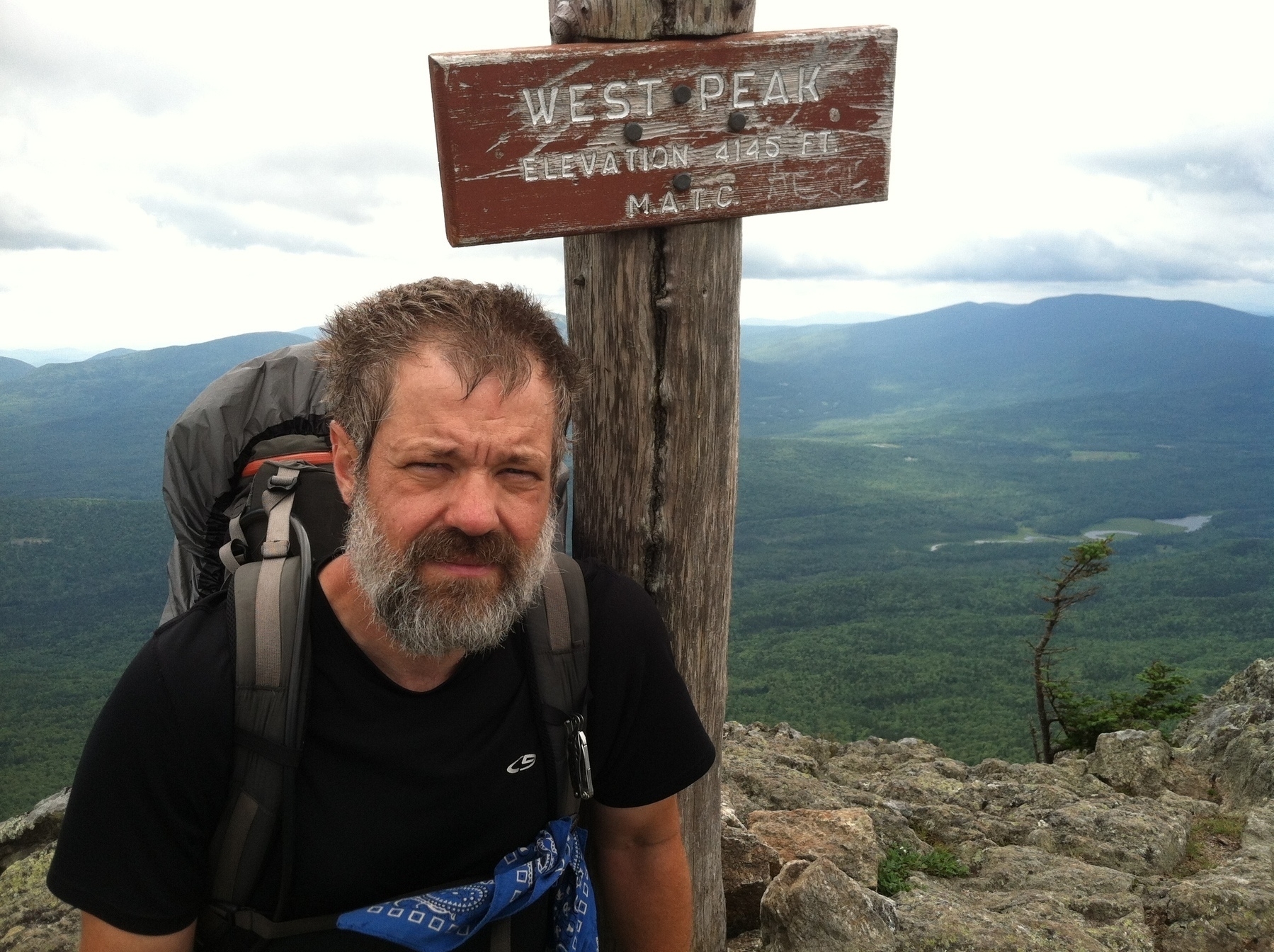 A man with a backpack is standing on a rocky mountain peak next to a wooden sign that reads "West Peak, Elevation 4145 FT, M.A.T.C.," with expansive forested hills in the background.