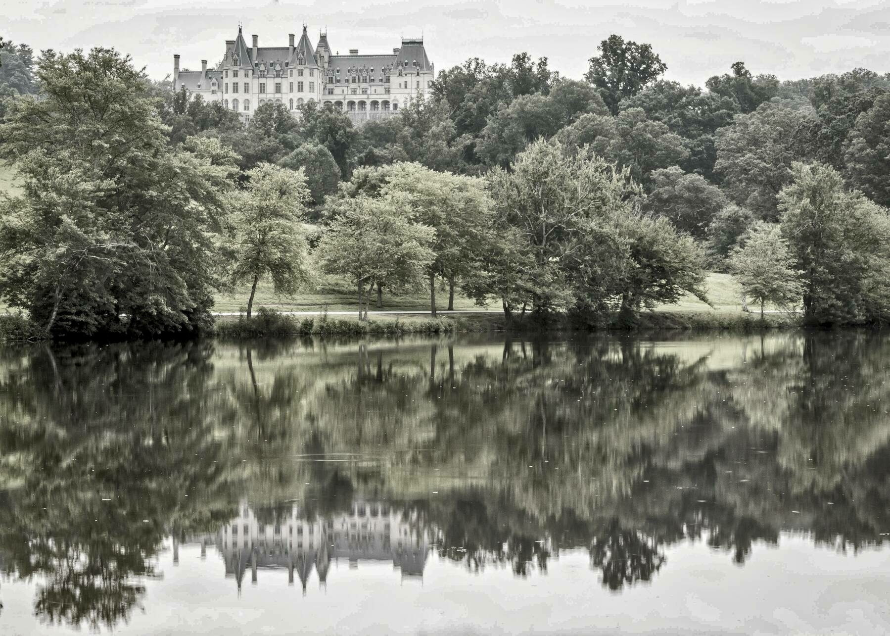 A large castle-like estate is visible in the background, perched atop a hill. In the foreground, a serene lake reflects the lush green trees lining its shore. The entire scene is enveloped in a peaceful, overcast ambiance, with gentle ripples on the water's surface mirroring the landscape above.