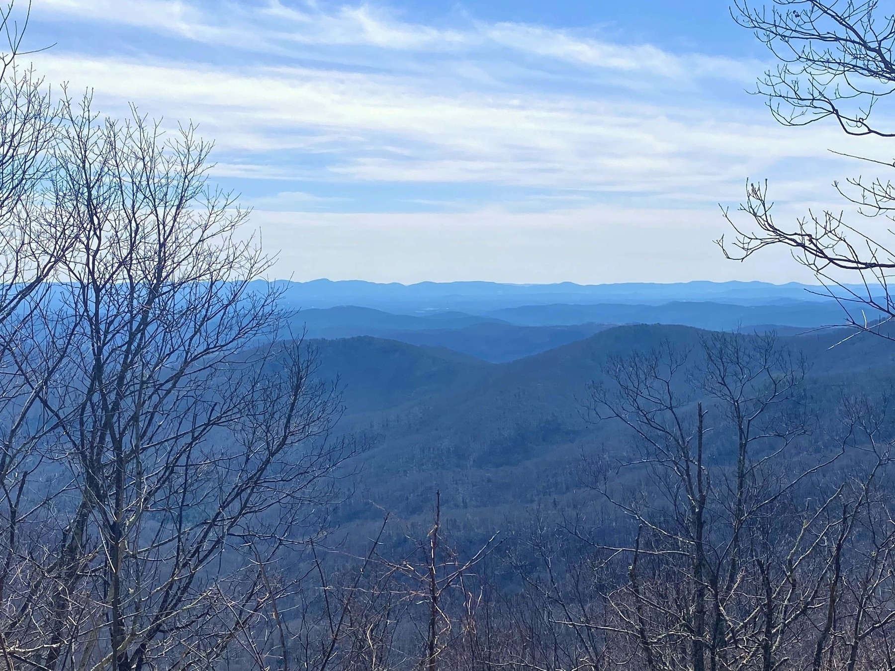 A scenic view of a mountainous landscape under a blue sky with some clouds. In the foreground, bare branches of trees are visible, framing the distant rolling hills and ridges that recede into the horizon. The mountains appear blue due to the atmospheric perspective, giving a sense of depth and vastness.